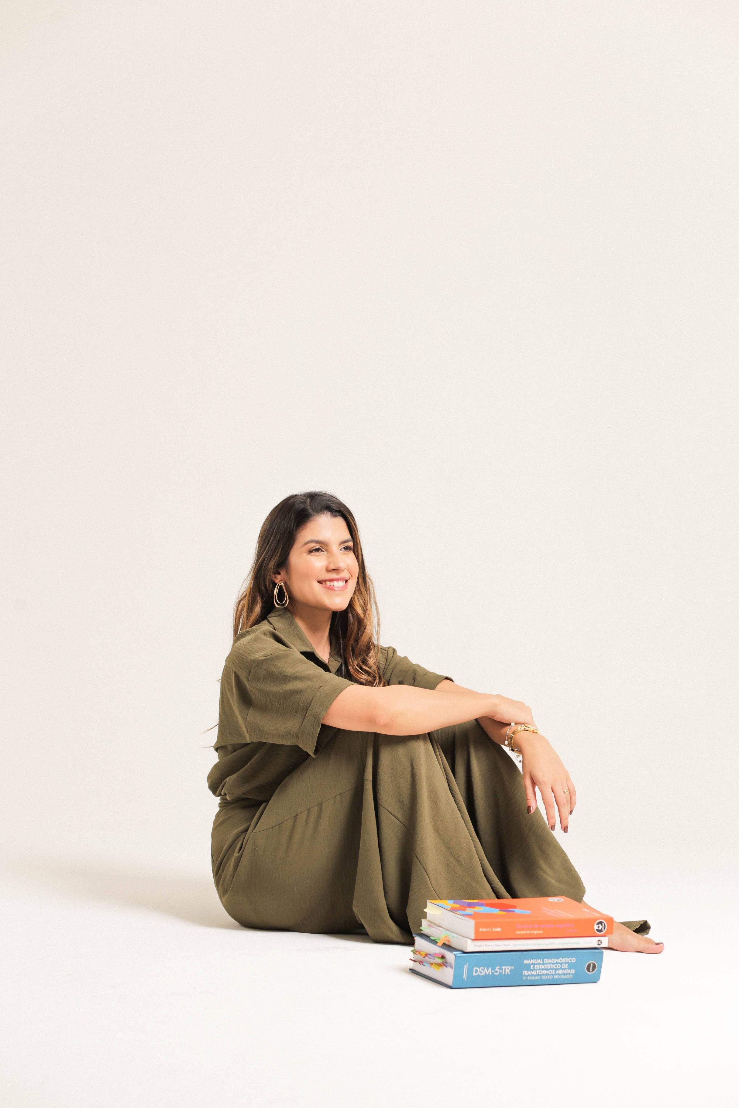 Natalie Cavalcante sentada no chão de um estúdio claro, sorrindo, ao lado de uma cadeira branca vazia.